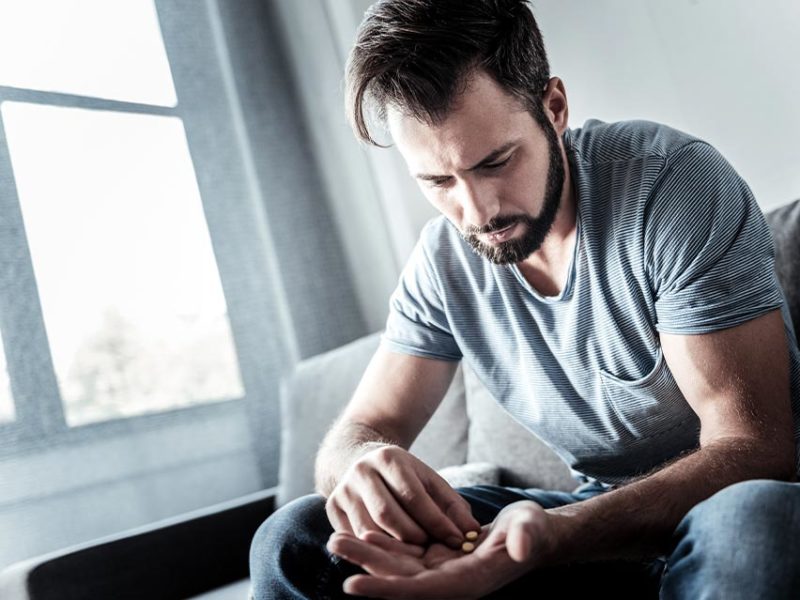distraught man showing signs of drug addiction sitting on a couch holding prescription pills