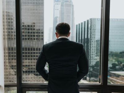 A well-dressed business man overlooking a city through his office window up high.