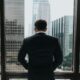 A well-dressed business man overlooking a city through his office window up high.