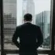 A well-dressed business man overlooking a city through his office window up high.