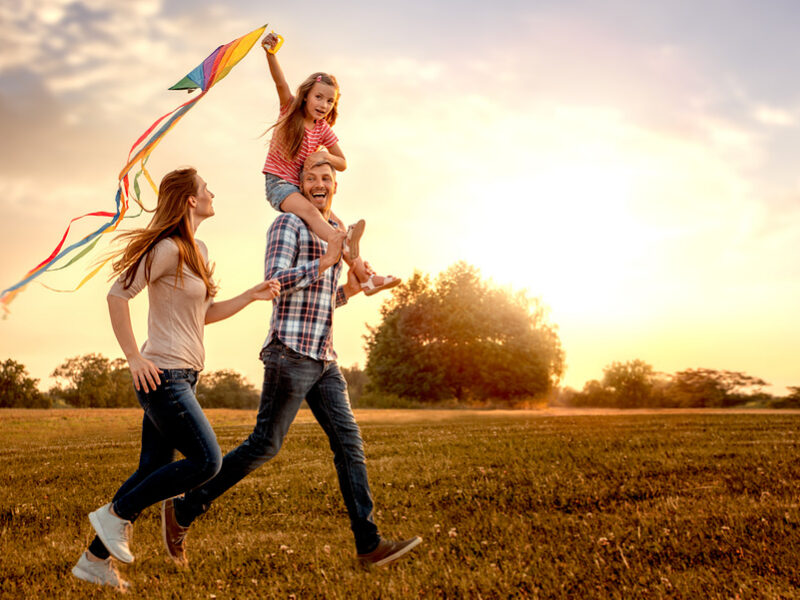 Happy family flying a kite together in a sunlit field at sunset, child on father's shoulders.