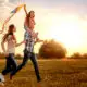 Happy family flying a kite together in a sunlit field at sunset, child on father's shoulders.