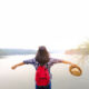 Woman with red backpack standing by a lake with arms outstretched, holding a hat, embracing freedom in recovery.
