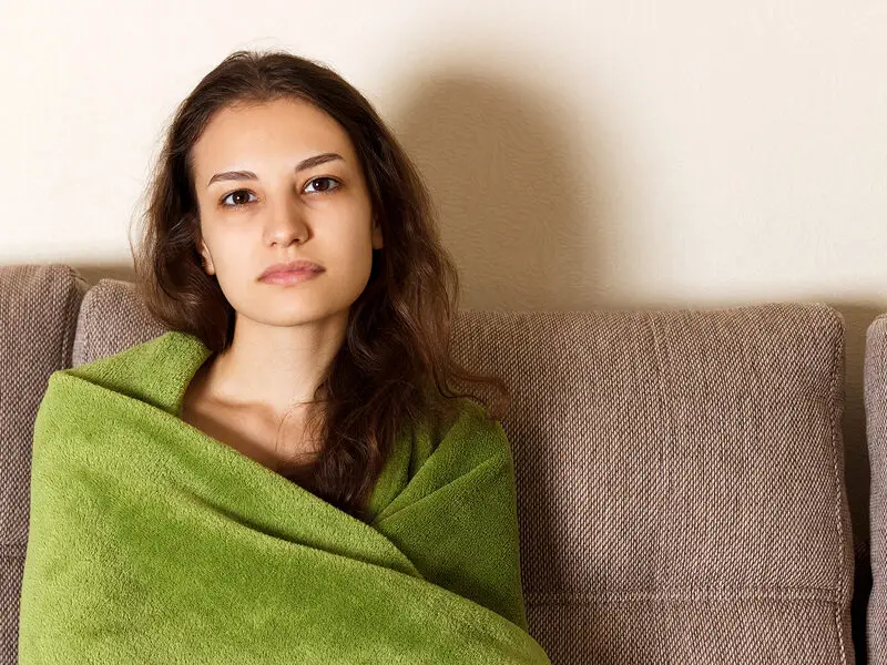 Young woman wrapped in a green blanket sitting on a couch, looking quietly reflective.