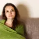Young woman wrapped in a green blanket sitting on a couch, looking quietly reflective.