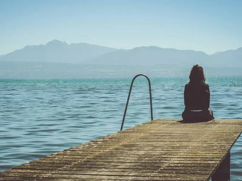 Woman sitting alone at the end of a wooden dock looking out over calm water toward distant mountains.