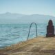 Woman sitting alone at the end of a wooden dock looking out over calm water toward distant mountains.