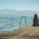 Woman sitting alone at the end of a wooden dock looking out over calm water toward distant mountains.