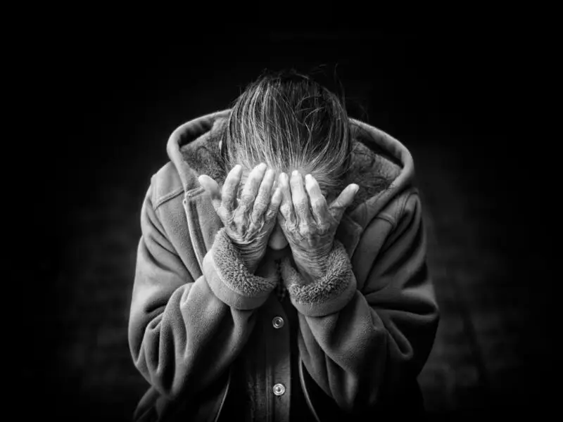 Black and white photo of an older person in a hooded coat covering their face with both hands in distress.