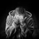 Black and white photo of an older person in a hooded coat covering their face with both hands in distress.