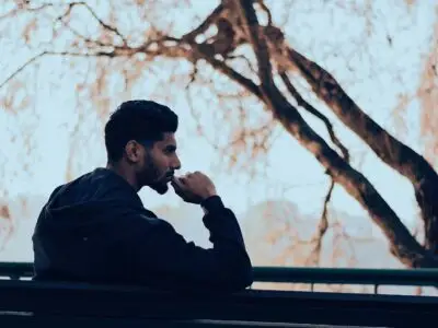 Man in a hoodie sitting outdoors by a railing, looking thoughtfully into the distance near bare trees.