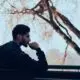 Man in a hoodie sitting outdoors by a railing, looking thoughtfully into the distance near bare trees.