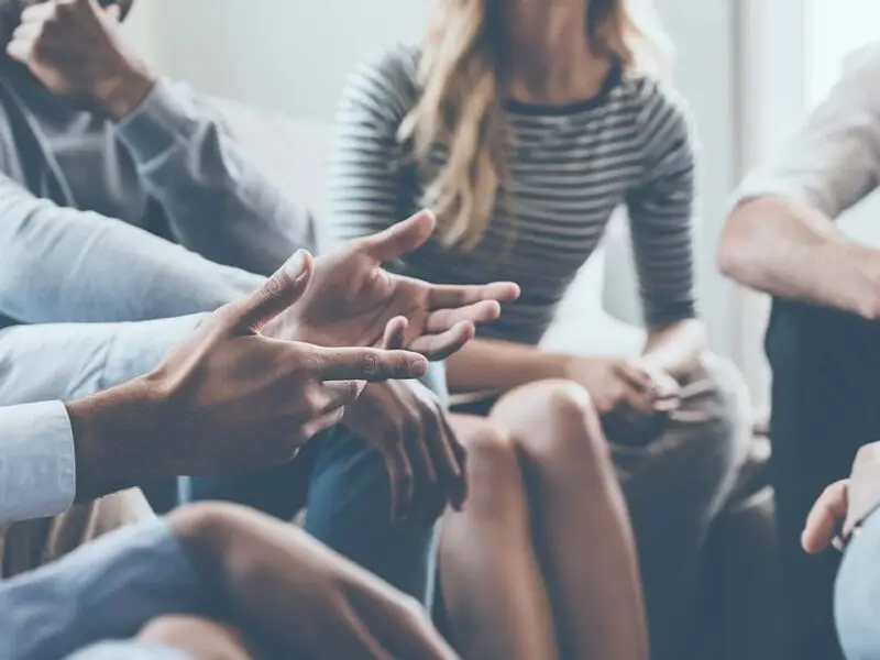Group therapy session with people seated in a circle, one person gesturing while speaking during discussion.