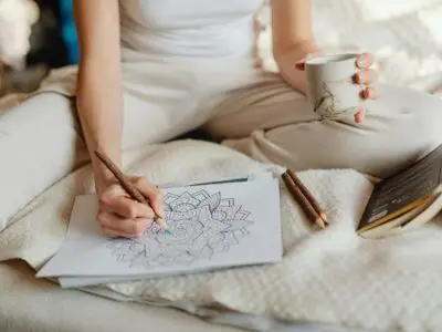 Person sitting on a bed coloring a mandala with colored pencils while holding a mug, a calming relaxation activity.