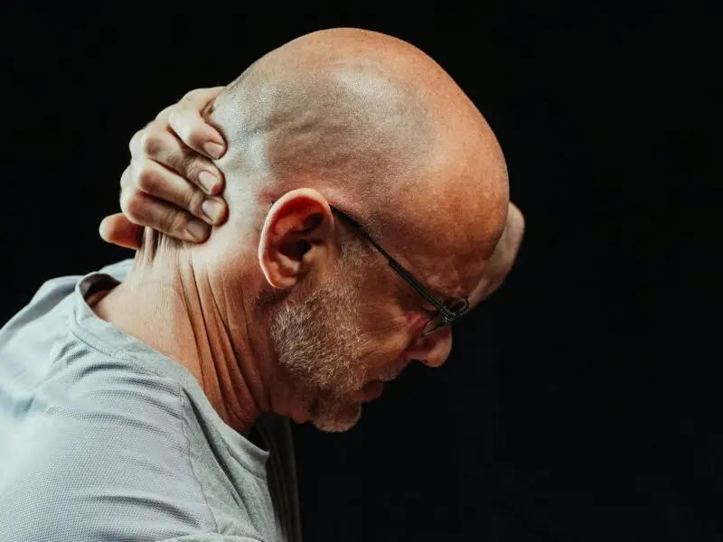Older bald man with glasses gripping his neck in pain, head bowed against a dark background.