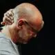 Older bald man with glasses gripping his neck in pain, head bowed against a dark background.