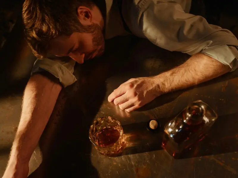 Man passed out at a table beside a glass and bottle of liquor, struggling with alcohol addiction.