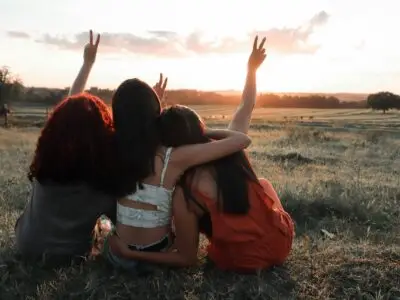 Three women sit together in a field at sunset, raising peace signs toward the sky, sharing a hopeful moment of connection.
