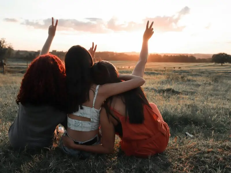Three women sit together in a field at sunset, raising peace signs toward the sky, sharing a hopeful moment of connection.