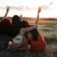 Three women sit together in a field at sunset, raising peace signs toward the sky, sharing a hopeful moment of connection.