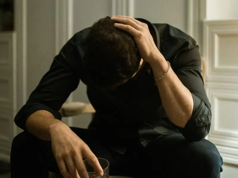 Man sitting with head in hand holding a glass of alcohol, struggling with addiction.