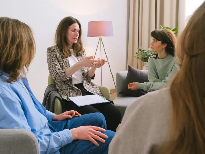 Therapist with clipboard leading a group therapy session with participants seated in a warm, homelike room.