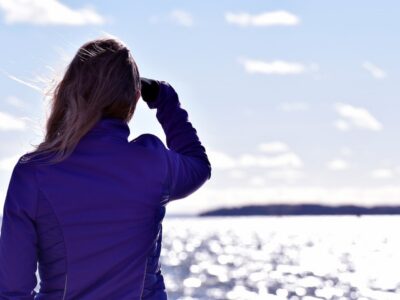 Woman in blue jacket looking out over sunlit water with binoculars, gazing toward the horizon.