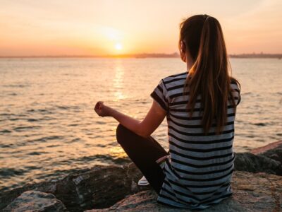 Woman meditating on rocks by the water at sunset along the Gulf Coast, finding calm during addiction recovery.