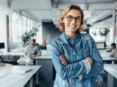 Smiling woman in denim jacket and glasses standing with arms crossed in a busy office with coworkers behind her.