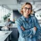 Smiling woman in denim jacket and glasses standing with arms crossed in a busy office with coworkers behind her.