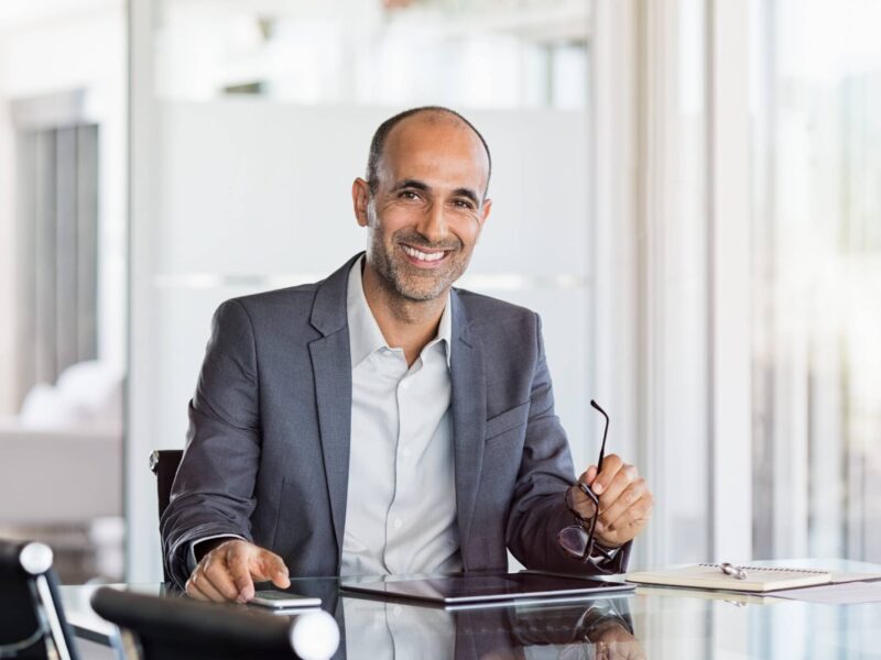 Smiling man in gray blazer sitting at a glass desk holding glasses, with a tablet and notebook nearby.