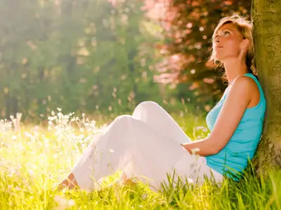 Woman relaxing against a tree in a sunlit meadow, reflecting peacefully during addiction recovery.