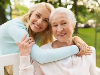 Smiling woman embracing an older woman outdoors on a bench, sharing a warm, supportive moment together.