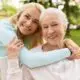 Smiling woman embracing an older woman outdoors on a bench, sharing a warm, supportive moment together.
