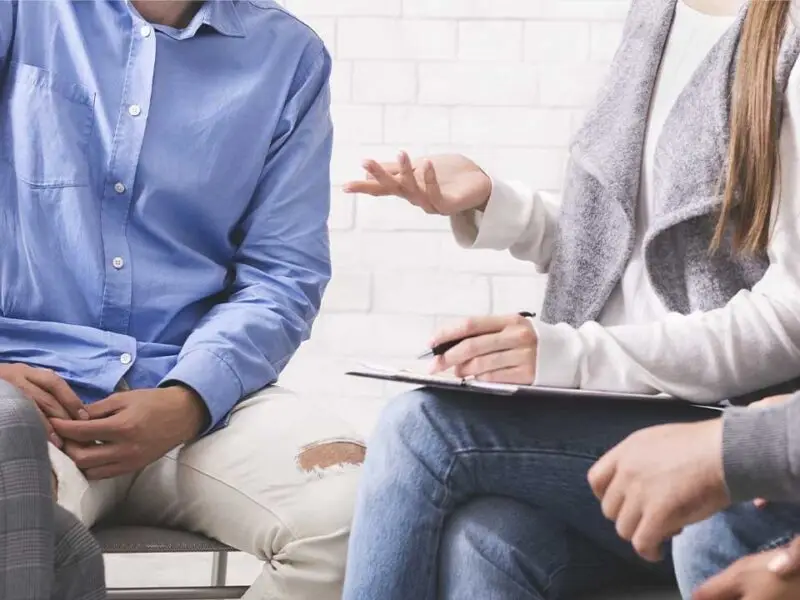 Counselor with clipboard leading a group therapy session with participants seated together during addiction treatment.