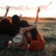 Three friends sit together in a field at sunset, arms raised in peace signs, sharing a supportive moment.