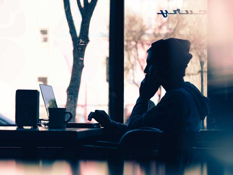 Silhouette of a person talking on the phone beside a laptop and coffee cup near a sunlit window.