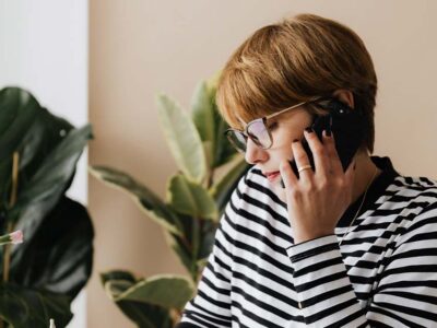 Woman in striped shirt and glasses making a phone call beside a leafy green plant.