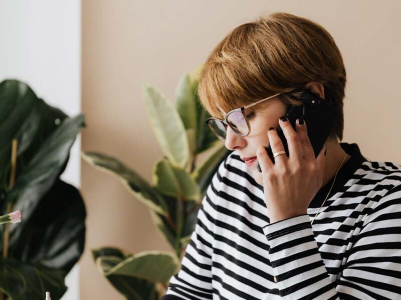 Woman in striped shirt and glasses making a phone call beside a leafy green plant.
