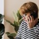 Woman in striped shirt and glasses making a phone call beside a leafy green plant.