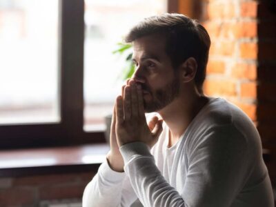 Thoughtful man sitting by a window with hands clasped, reflecting on seeking addiction treatment and recovery.
