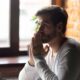 Thoughtful man sitting by a window with hands clasped, reflecting on seeking addiction treatment and recovery.