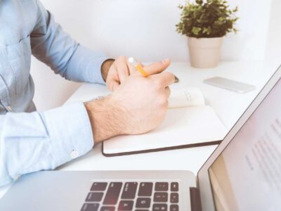 Person writing in a notebook with a pencil beside a laptop at a bright desk with a small potted plant.
